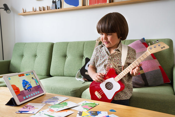 Child playing a red Loog Mini guitar beside a sage sofa and music learning cards.