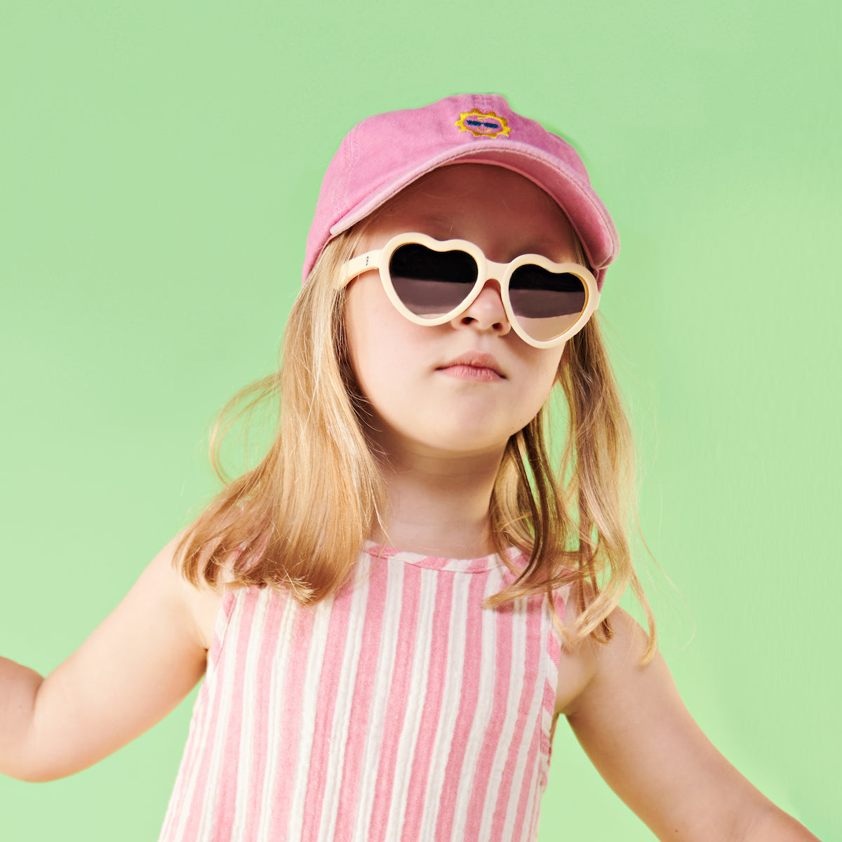 Child wearing cream heart-shaped Babiators sunglasses with a pink cap against a mint backdrop.